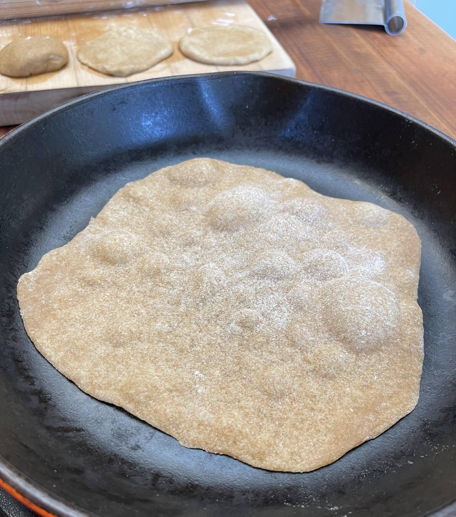 Bubbles form on the surface of a wheat tortilla cooking in an enameled cast iron pan. More tortillas sit in the background, ready to roll out and cook.