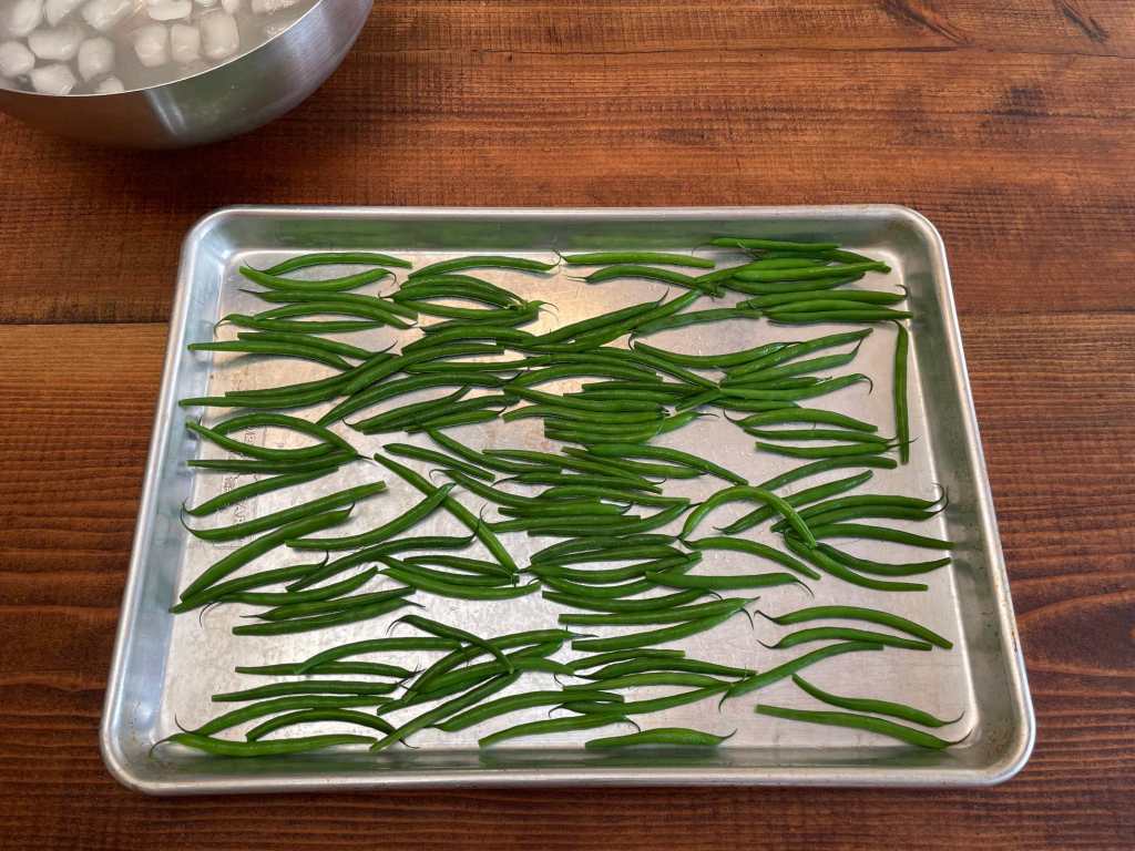 Blanched green beans arranged in a single layer on a silver cookie sheet that sits on a dark wooden table. A stainless steel bowl of ice water sits in the top left.