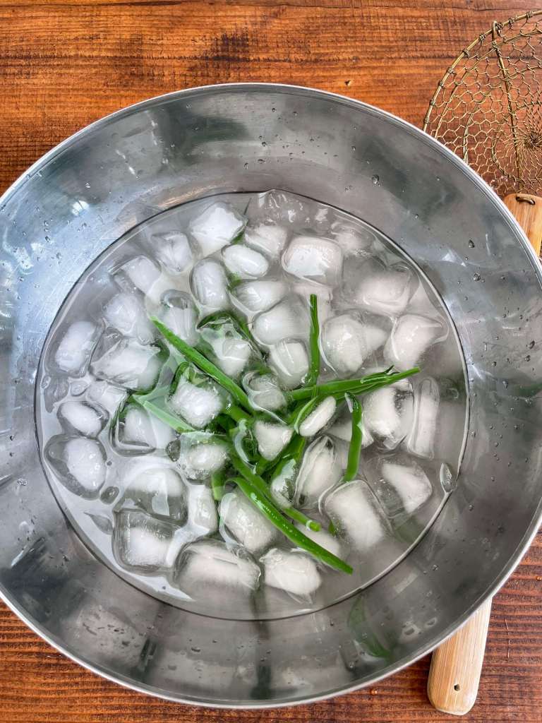 Blanched green beans cool in a large stainless steel bowl of water with ice cubes