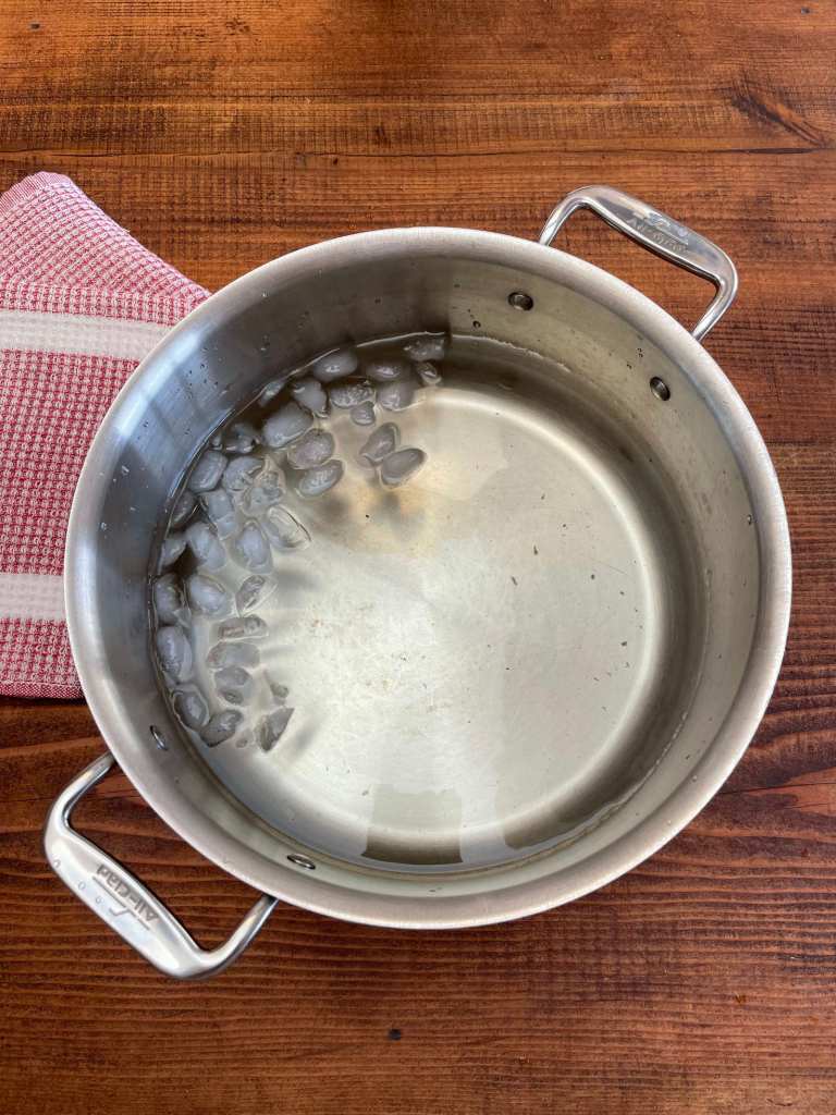 A large stainless steel pot of water with small ice cubes off to the left side. The pot sits on a dark wood table. A red checked dishtowel sits to its left.