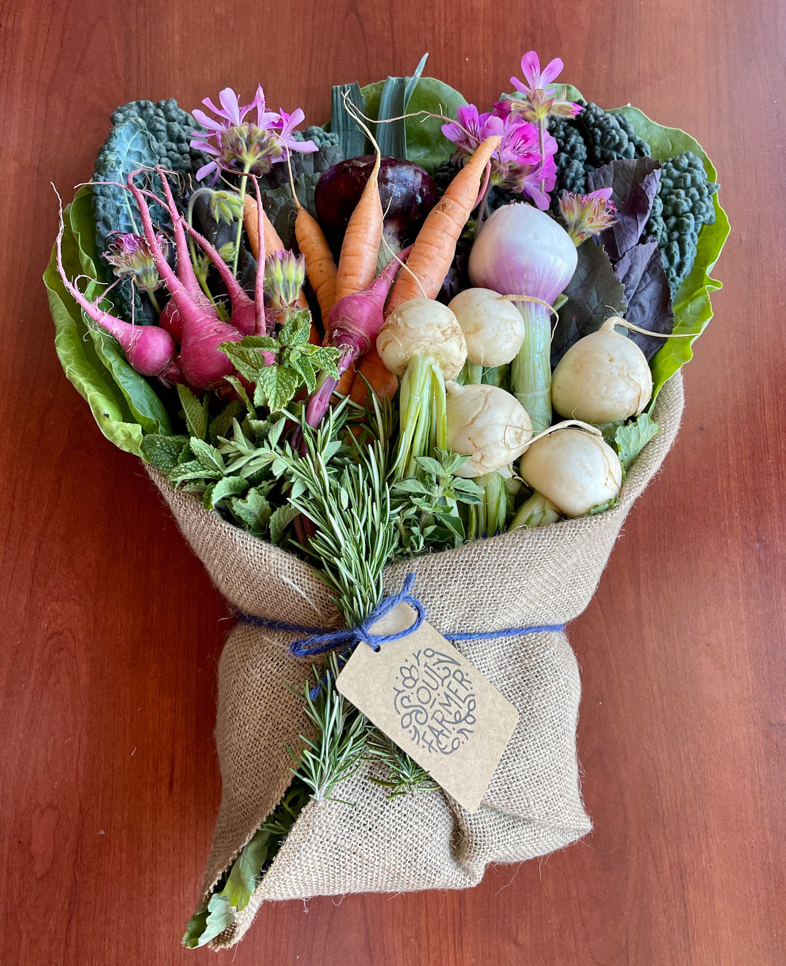 A bouquet of fresh vegetables wrapped in burlap sits on a wooden background