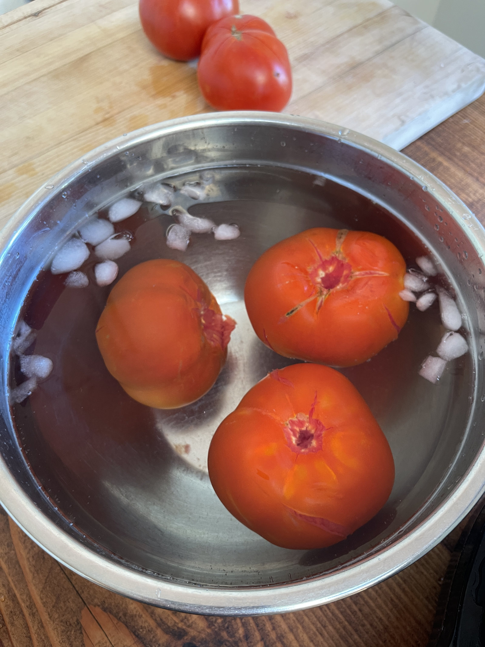 Three large blanched tomatoes cool in a stainless steel bowl filed with ice water and melting ice cubes. In the background is a cutting board with two fresh tomatoes sitting on it.
