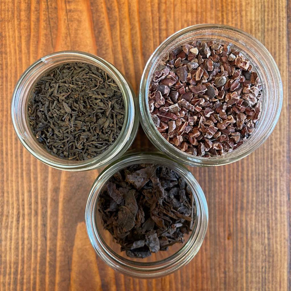 Three jars sitting on a wooden background. Two contain puerh tea. The top right jar contains cacao nibs.