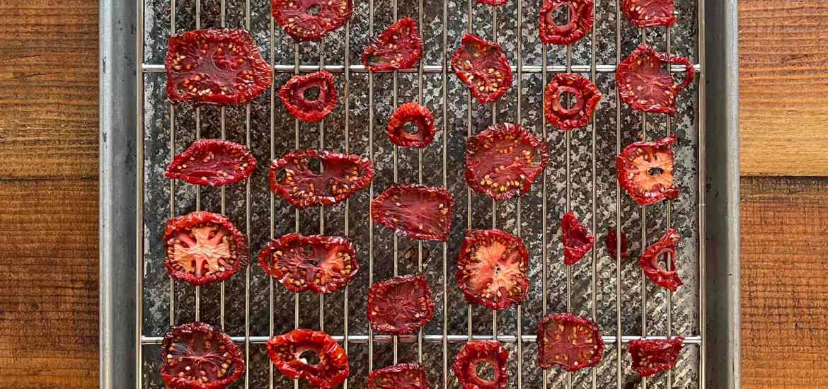 Dehydrated tomato slices arranged on a silver wire cooling rack set inside a silver baking sheet before dehydrating. The baking sheet sits on a dark wood background.