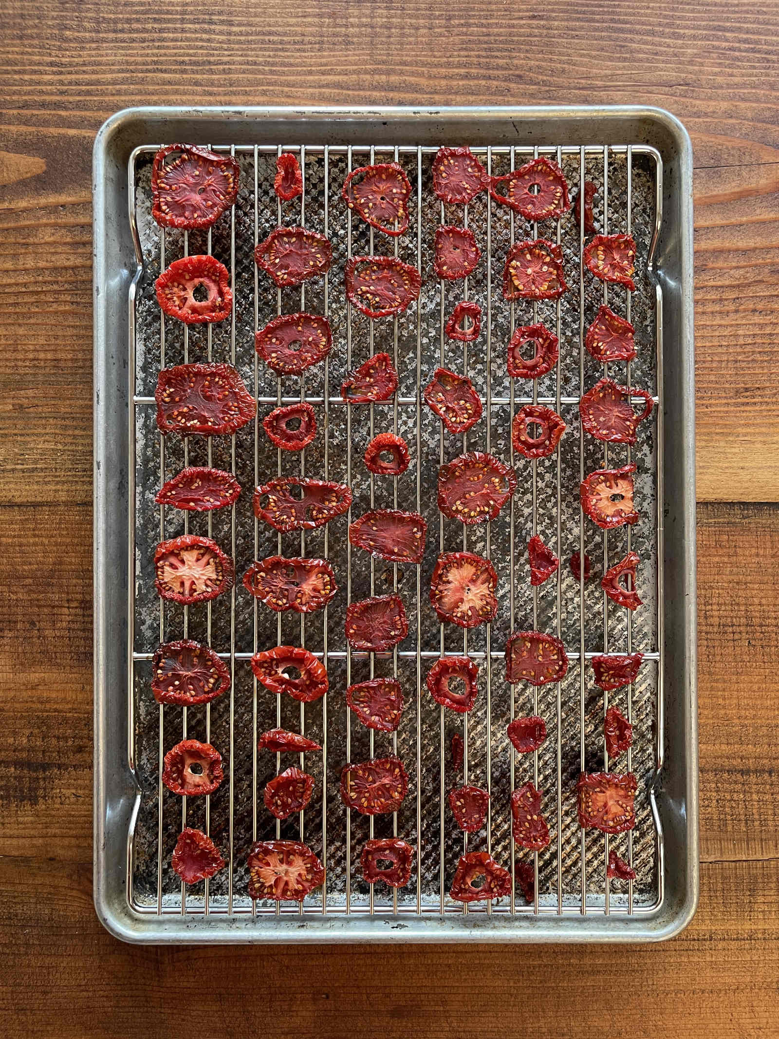 Dehydrated tomato slices arranged on a silver wire cooling rack set inside a silver baking sheet before dehydrating. The baking sheet sits on a dark wood background.