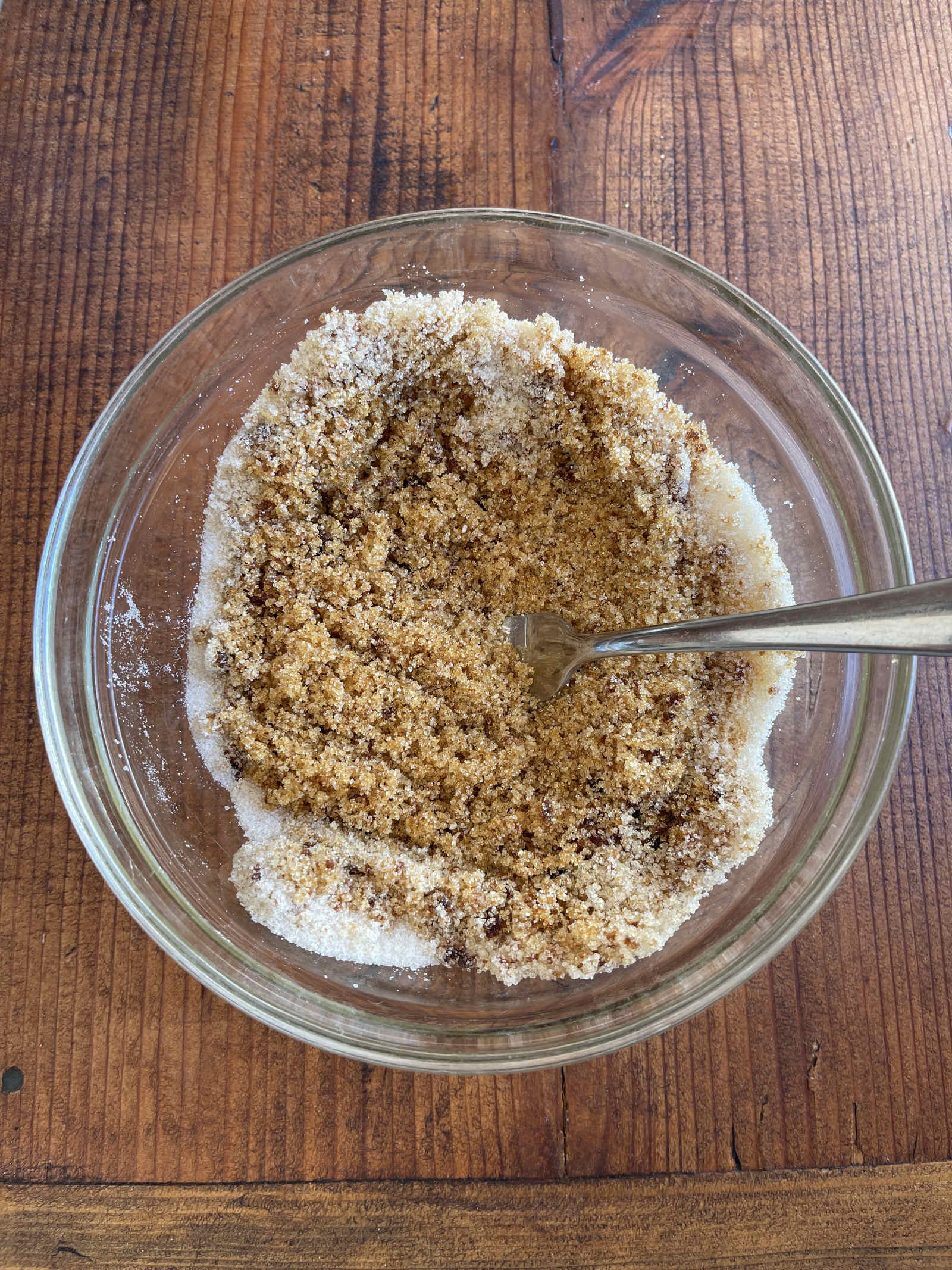 Partially mixed granulated sugar and molasses on their way to becoming brown sugar. The mixture sits in a glass bowl on a dark wooden tabletop.