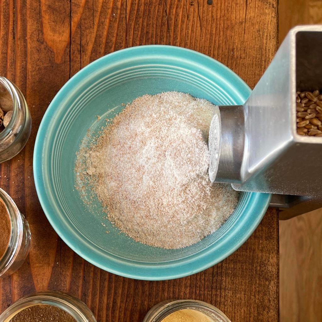 A turquoise bowl set below the grinder of a grain mill is filled with freshly ground whole wheat flour