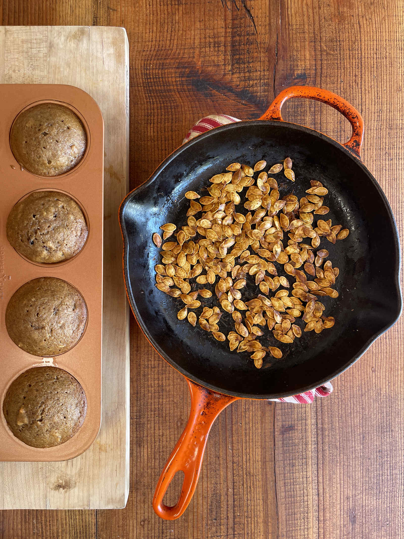 A cast iron pan filled with roasted pumpkin seeds sits on the right of a partially shown muffin pan filled with pumpkin spice muffins