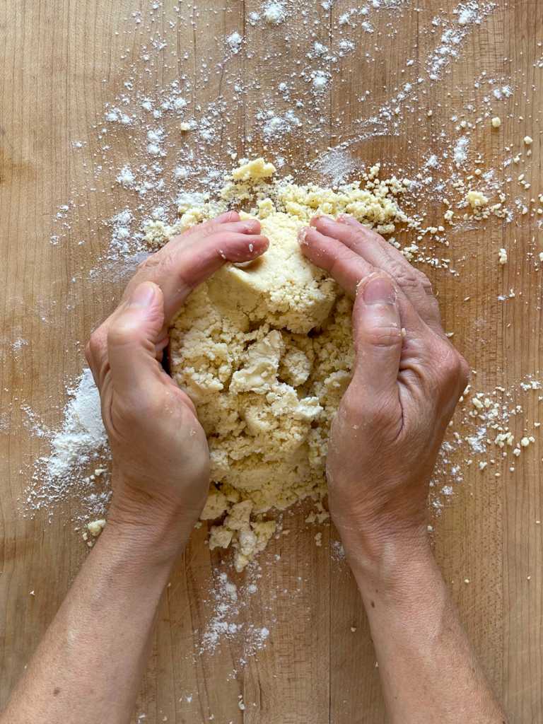 Hands are pushing freshly mixed semolina pastry together on a light colored wooden cutting board dusted with flour