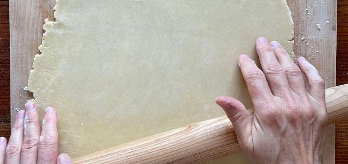 Hands roll out semolina pastry using a tapered rolling pin on a lightly flour-dusted wooden board