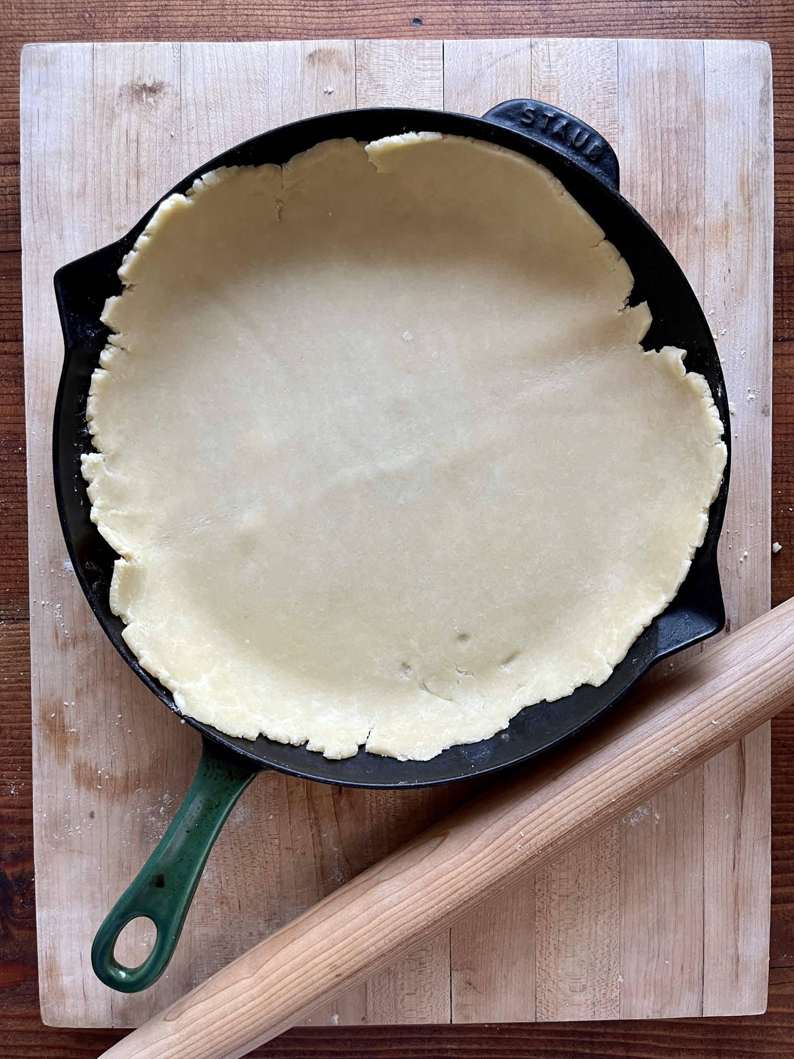 Semolina pastry lines a 12-inch cast iron pan. A tapered rolling pin rests below it. The background is a light colored wooden cutting board.