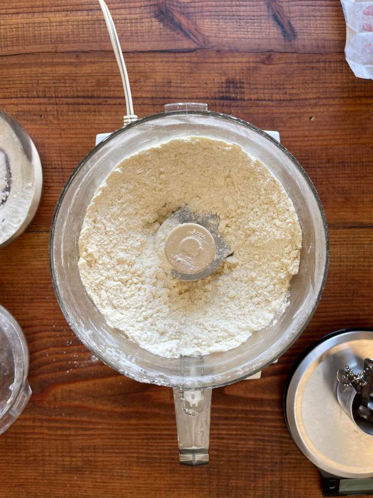 Flour and butter have been processed in a food processor to make pastry. The food processor sits on a dark wooden tabletop. A scale sits in the bottom right corner.