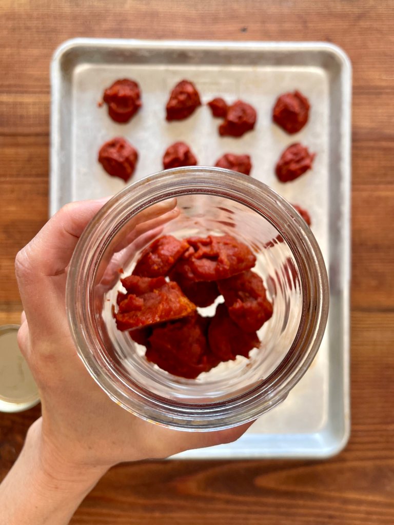 A jar is filling up with blobs of frozen tomato paste. A silver cookie sheet covered in blobs of frozen tomato paste sits on a wooden table in the background.