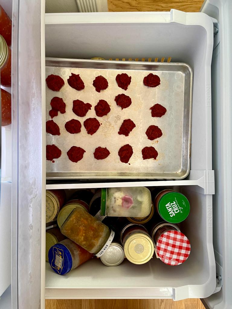 A silver cookie sheet in the freezer has frozen blobs of tomato paste spread across it. In the compartment next to the cookie sheet are a bunch of glass jars filled with frozen food.