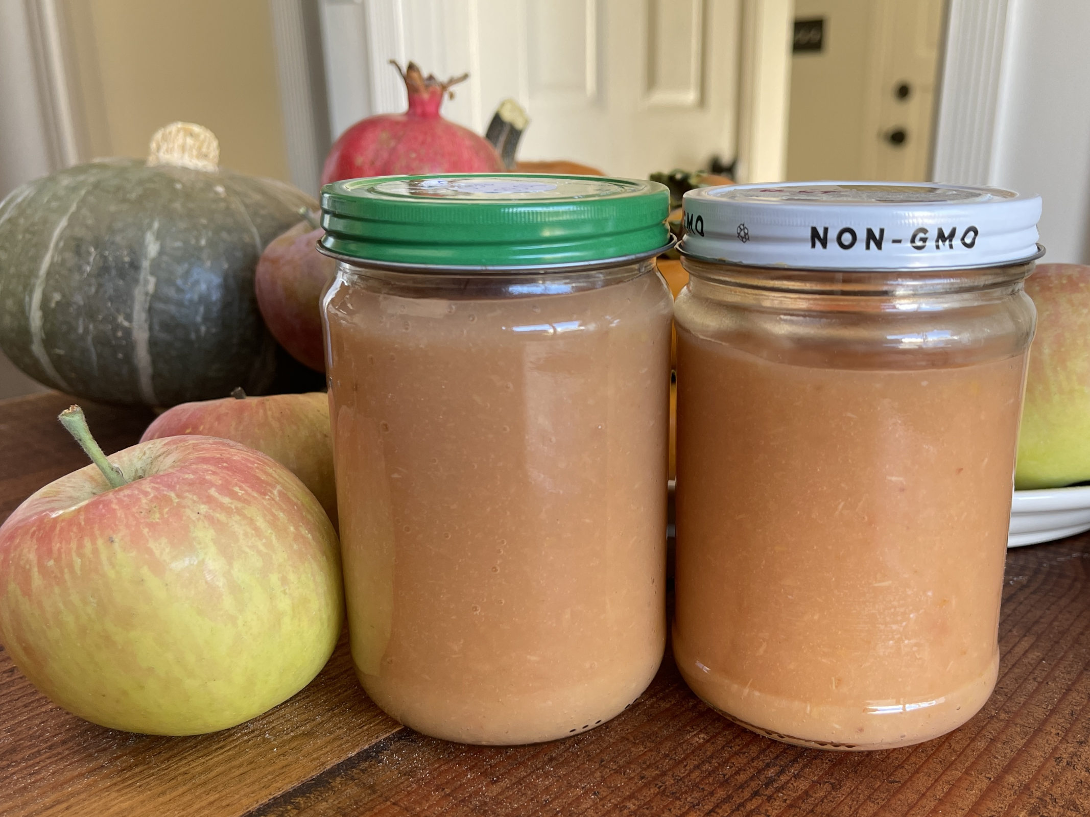 Two jars of applesauce sit on a wooden table, surrounded by fresh fall produce