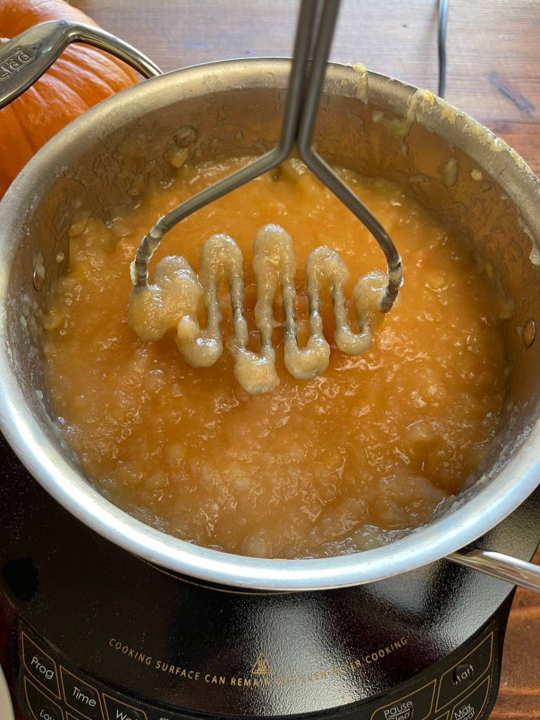 A potato masher is mashing cooked apple chunks down into chunky applesauce