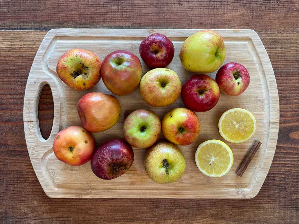 Apples, a halved lemon and a cinnamon stick sit on a light wooden cutting board