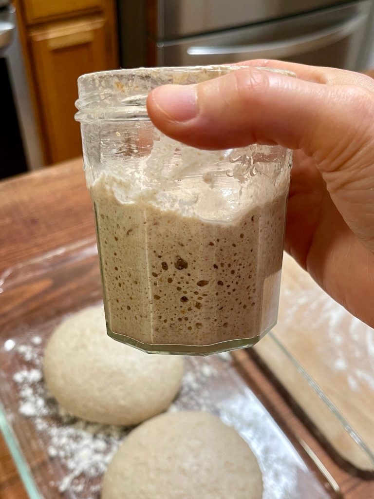 A hand holds a jar of active sourdough starter filled with bubbles. Two balls of pizza dough rise in the background.