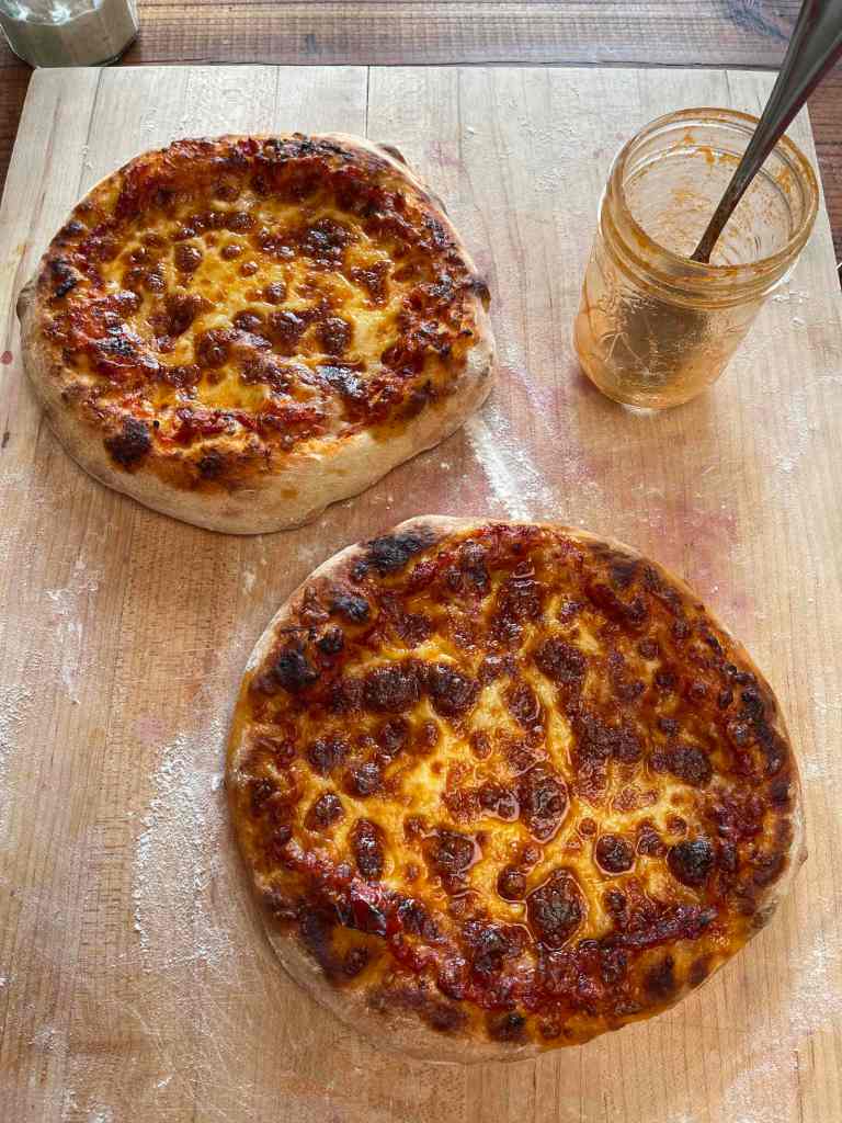 Two cheese sourdough pizzas cool on a wooden cutting board. An empty jar of pizza sauce sits in the top right with a spoon in it.