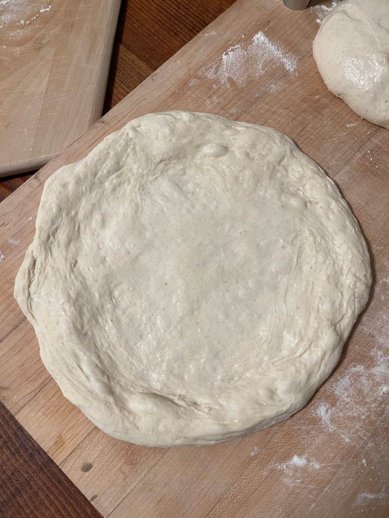 Formed sourdough pizza dough sits on a floured wooden board
