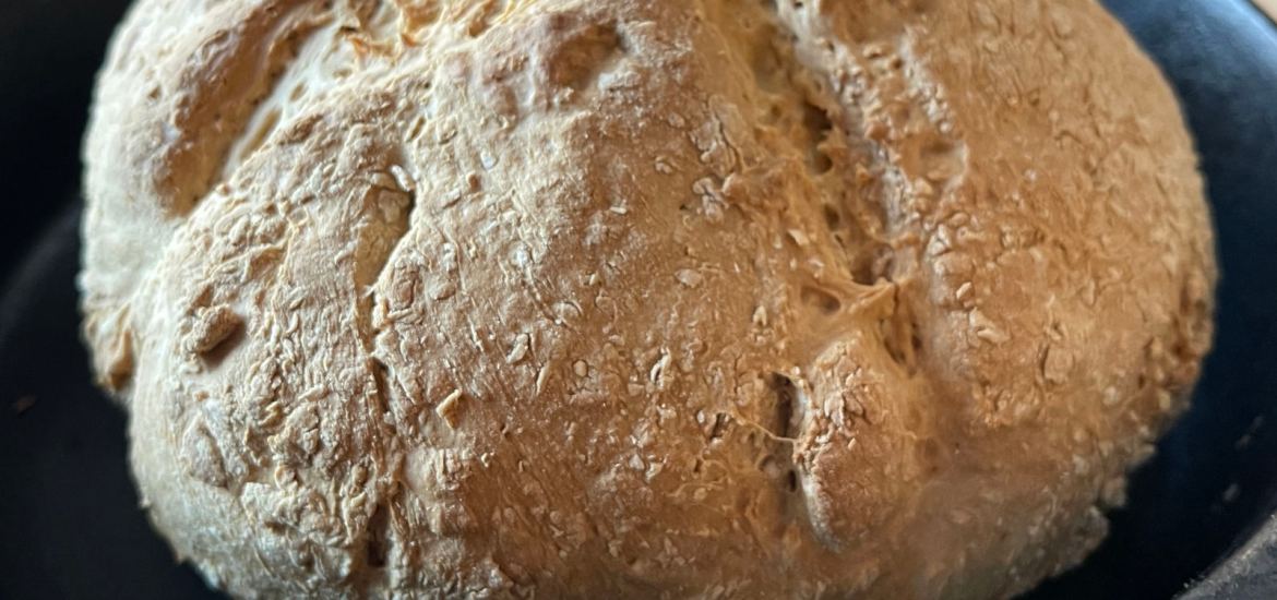 A hot loaf of freshly baked vegan soda bread baked with soy buttermilk cools in a cast iron pan sitting on a metal rack.