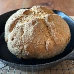 A hot loaf of freshly baked vegan soda bread baked with soy buttermilk cools in a cast iron pan sitting on a metal rack.