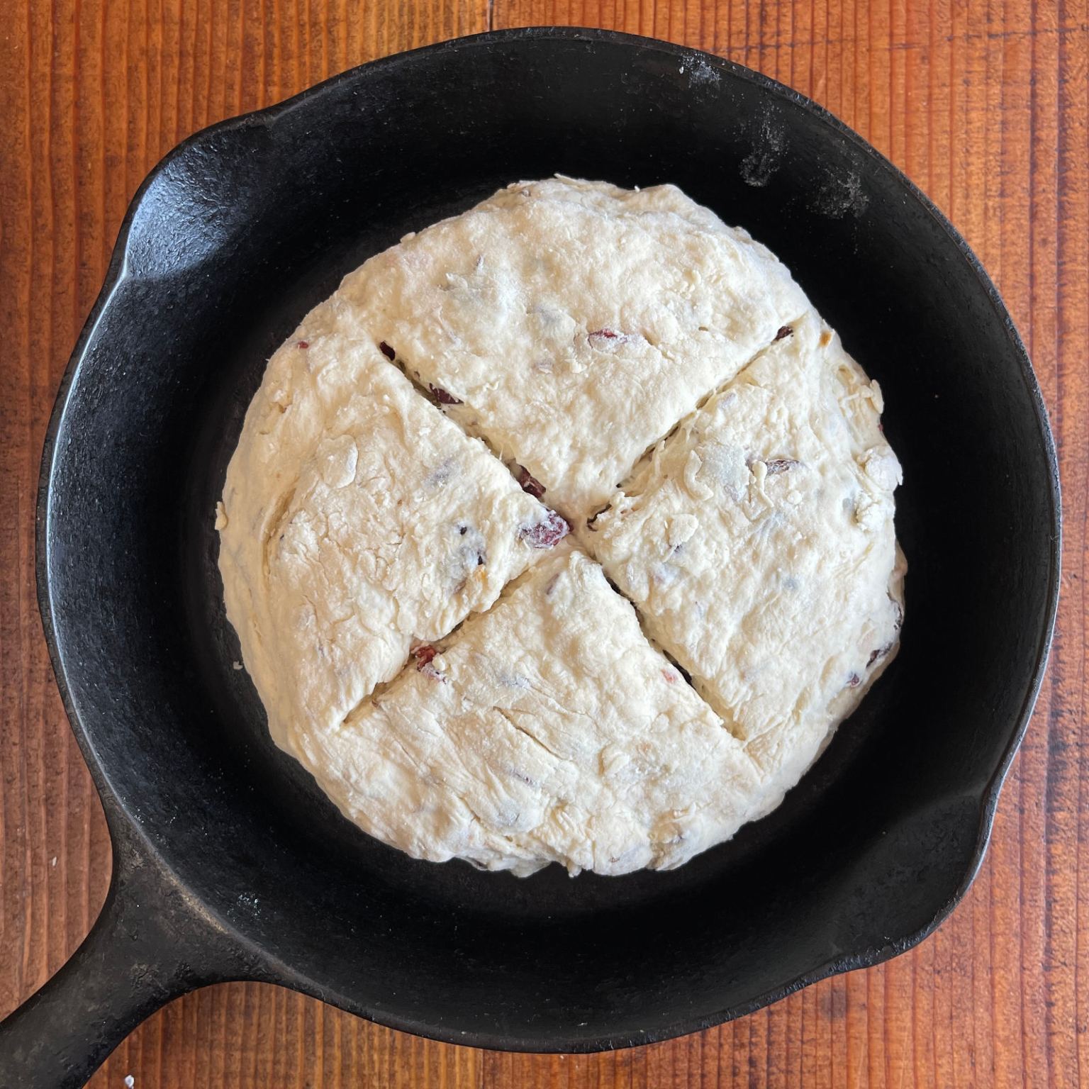A loaf of vegan cranberry soda bread is formed and scored and ready to be baked. It is in a cast iron pan.