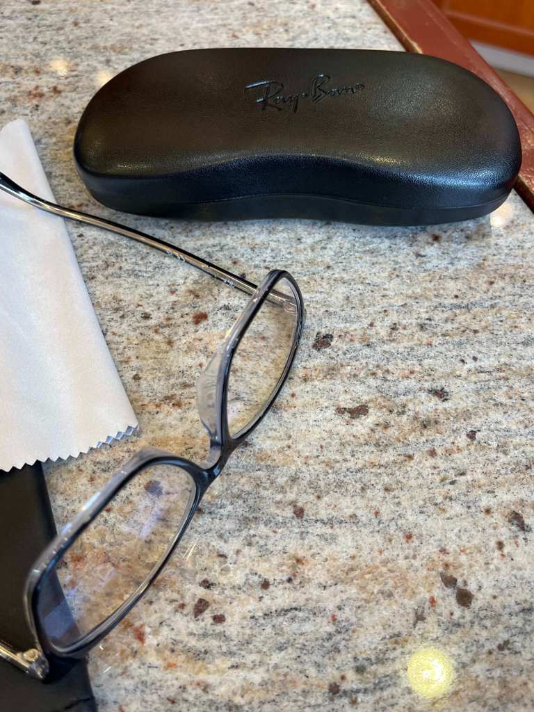 A pair of black framed glasses, a grey microfiber cleaning cloth and a black glasses case sit on a marble background