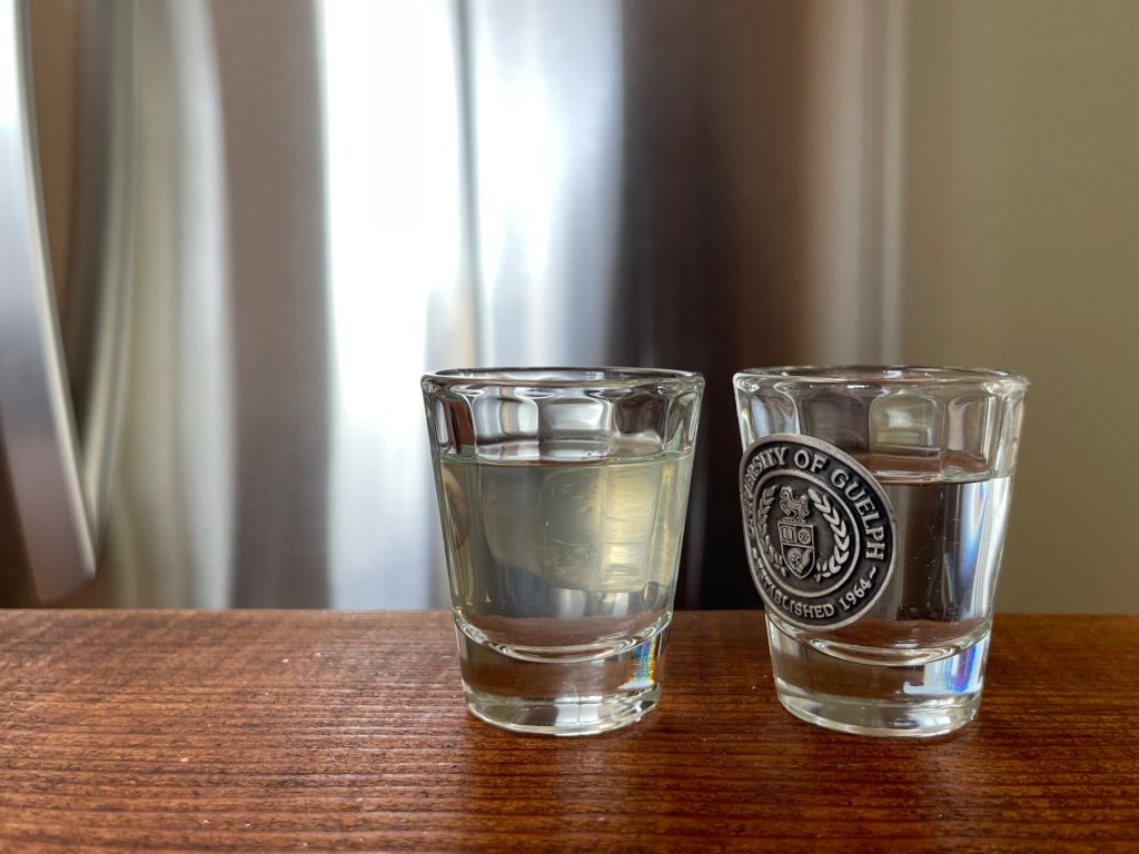 Two shot glasses sit on a dark wooden table. The one on the left is filled will pale yellow homemade vinegar. The one on the right is filled with water. 