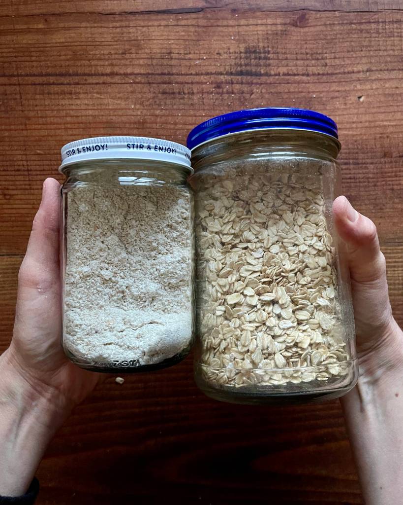 Hands hold a smaller jar of oat flour (left) and a larger flour of rolled oats (right)