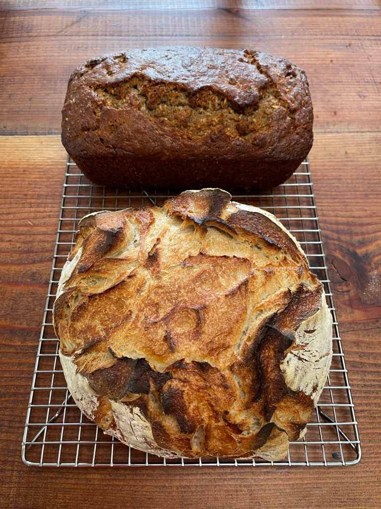 A loaf of sourdough bread sits in the foreground. A loaf of coconut banana nut quick bread sits behind it. The loaves are cooling on a silver wire rack sitting on a wooden table.