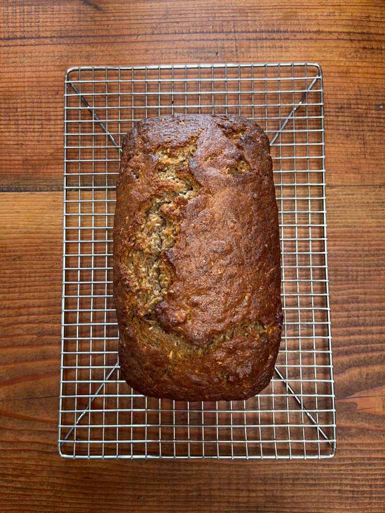 A loaf of coconut banana nut bread cools on a silver wire rack on a wooden background