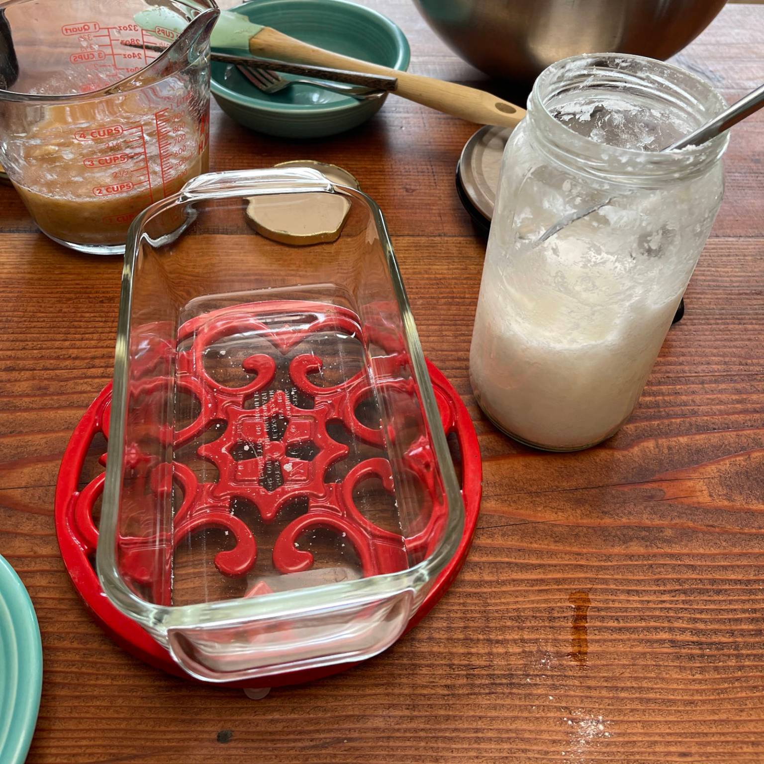 On a wooden table, melted coconut oil cools in a glass loaf pan sitting on a red metal trivet. A jar of solid, unmelted coconut oil sits beside the loaf pan.