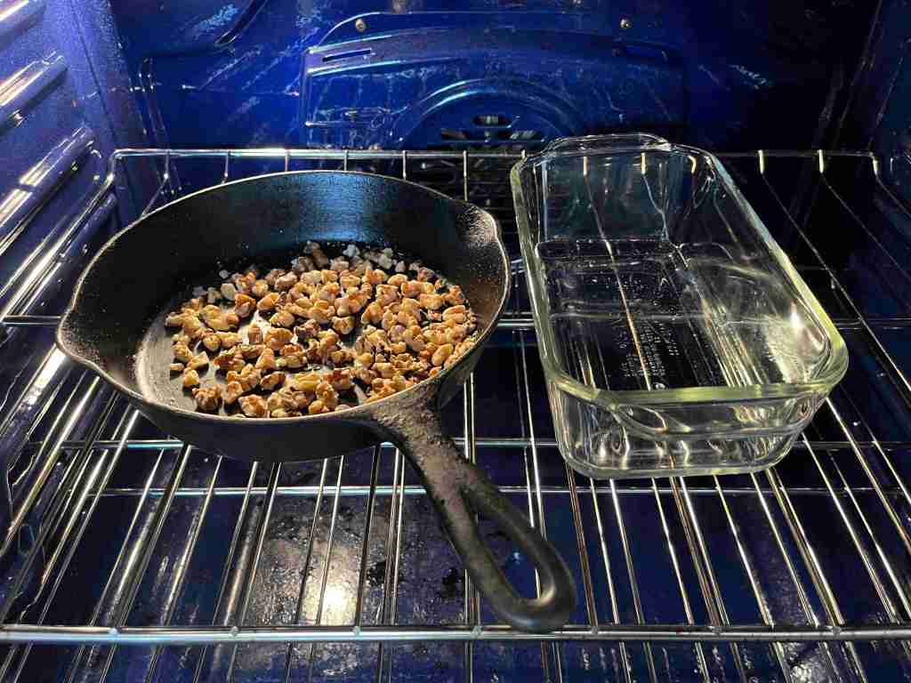 Inside an oven, walnuts roast in a cast iron pan. Coconut oil melts in a glass bread loaf pan.