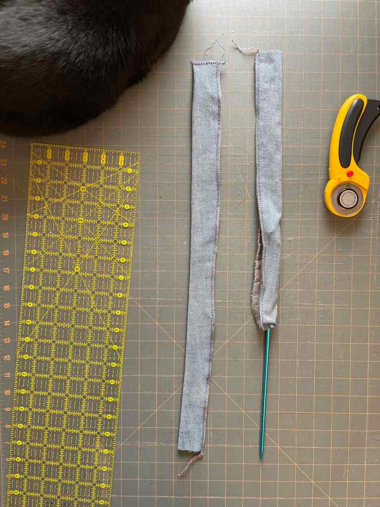 Two strips of denim have been sewn into tubes for bag handles. A knitting needle turns one tube inside out. The handles and sewing tools sit on a gray cutting mat. A black cat sits in the top left corner.