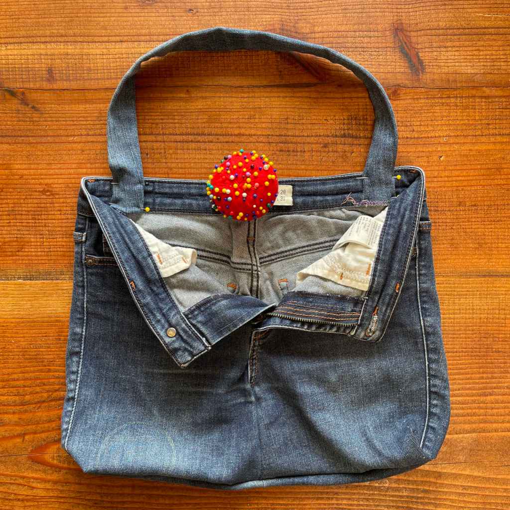 A handle has been pinned onto the inside back of a denim shopping bag made out of a pair of old jeans. A red pin cushion sits in the middle of the waist band.