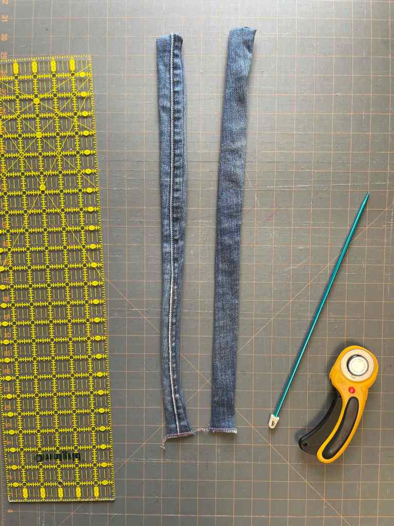 Two denim bag handles made for a denim bag sit on a gray cutting mat. Sewing tools sit around them.