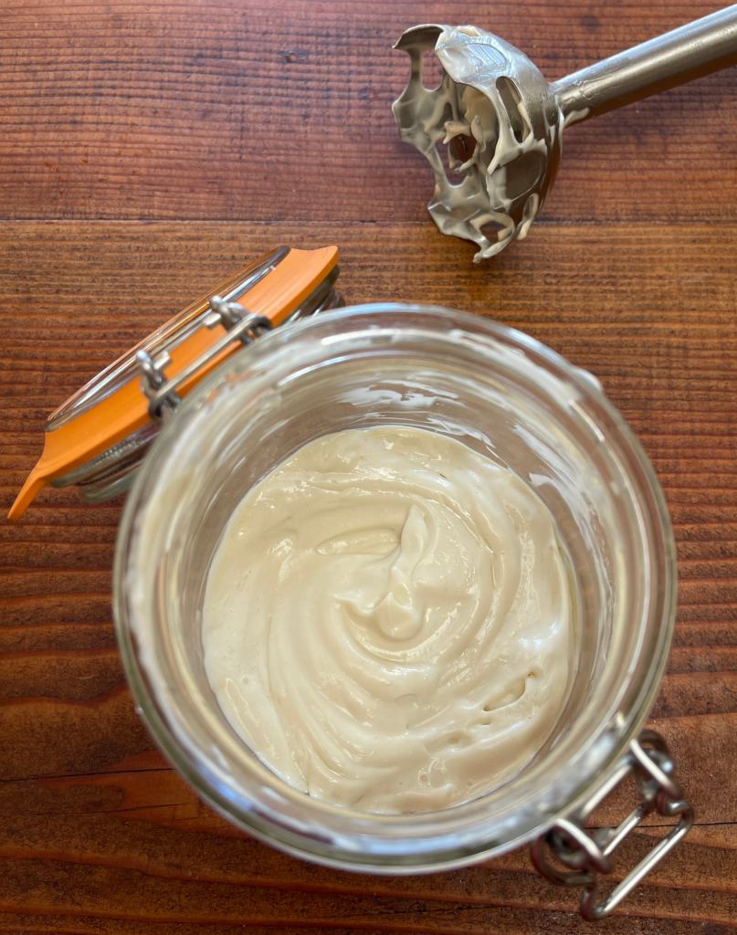 Mayonnaise made with aquafaba sits in a glass jar. Above it is the blade of a stainless steel immersion blender used to make the mayonnaise.
