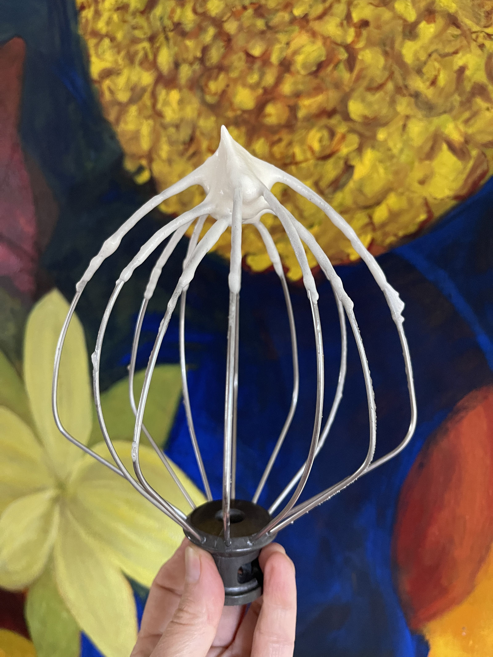 The wire whip attachment of a stand mixer has a stiff white peak of whipped aquafaba on the tip. In the background is a painting of yellow flowers.