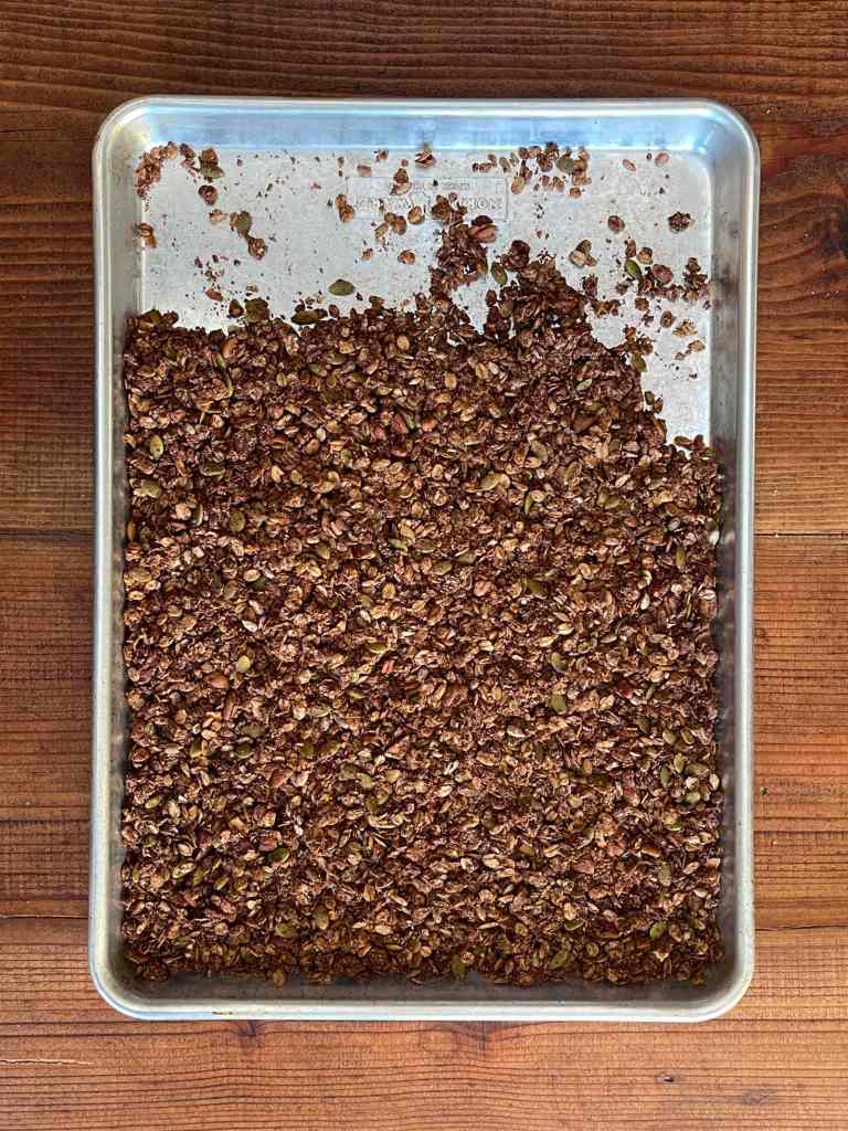 Baked chocolate and nut granola on a silver baking sheet. The baking sheet sits on a dark wood tabletop.