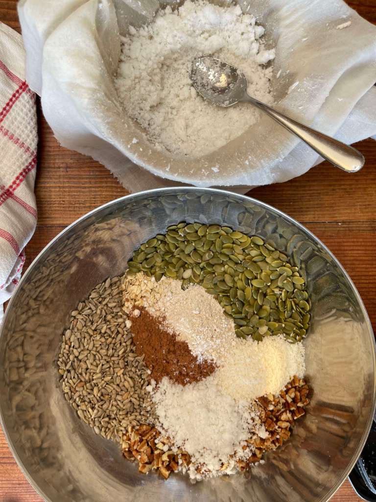 The dry granola ingredients are in a large stainless steel bowl. Above it sits a cloth-lined bowl that contains coconut pulp leftover from making coconut milk.