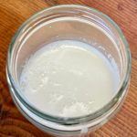 A glass jar half full with thick and somewhat frothy coconut milk. The jar sits on dark wood.