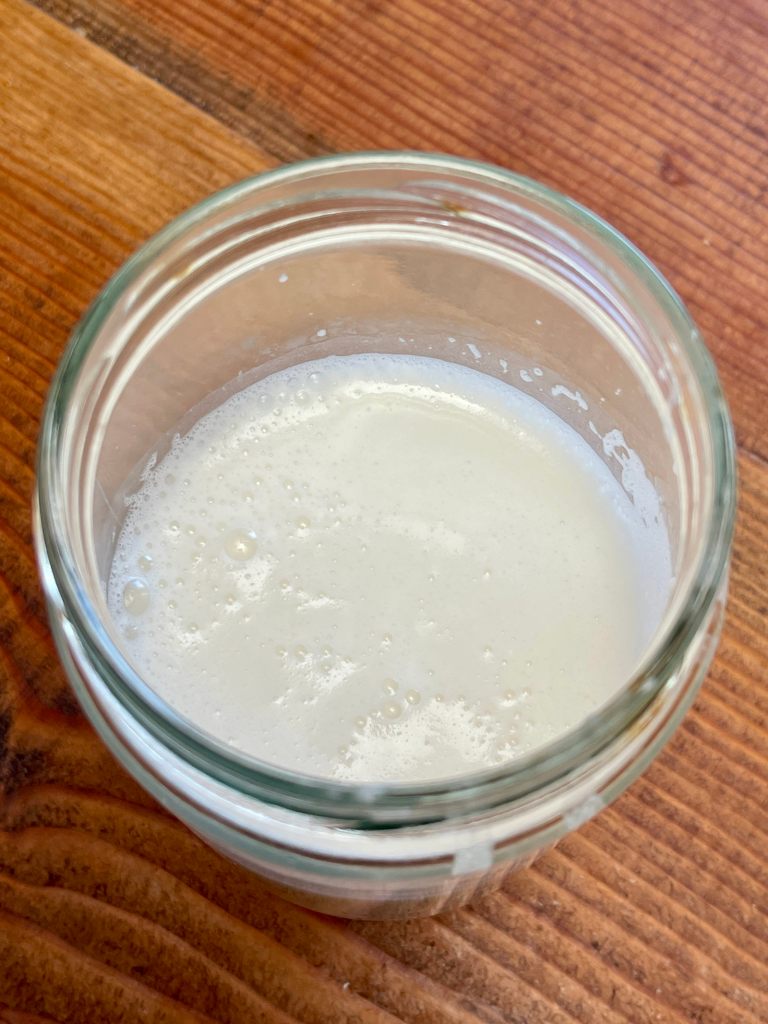 A glass jar half full with thick and somewhat frothy coconut milk. The jar sits on dark wood.
