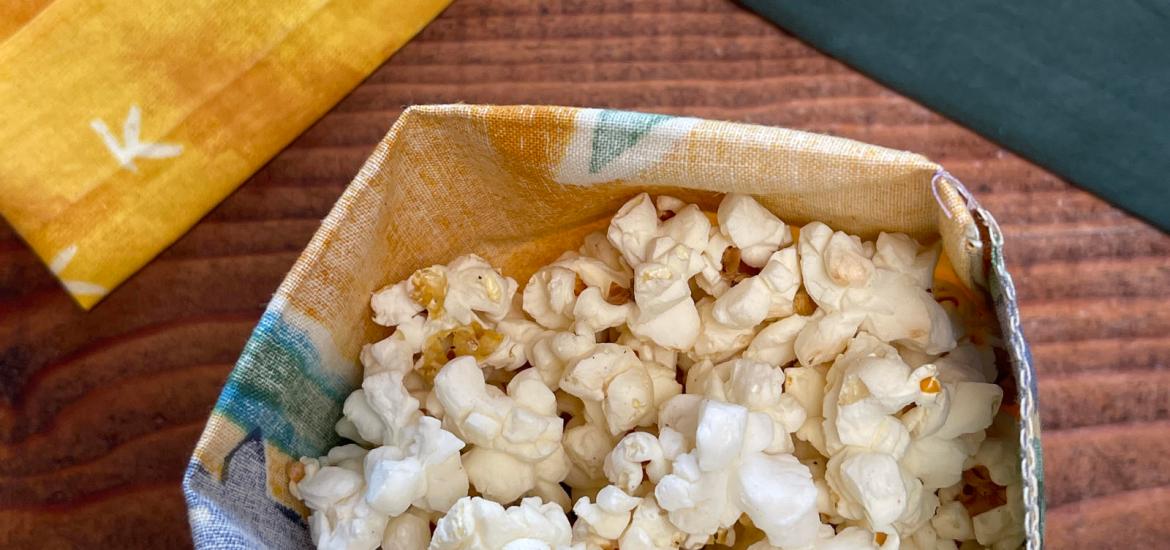 A homemade yellow, blue and green reusable cloth snack bag has been filled with popcorn. An empty yellow snack bag sits to the left. On the right is an empty dark green snack bag. Everything sits on a dark wooden table.