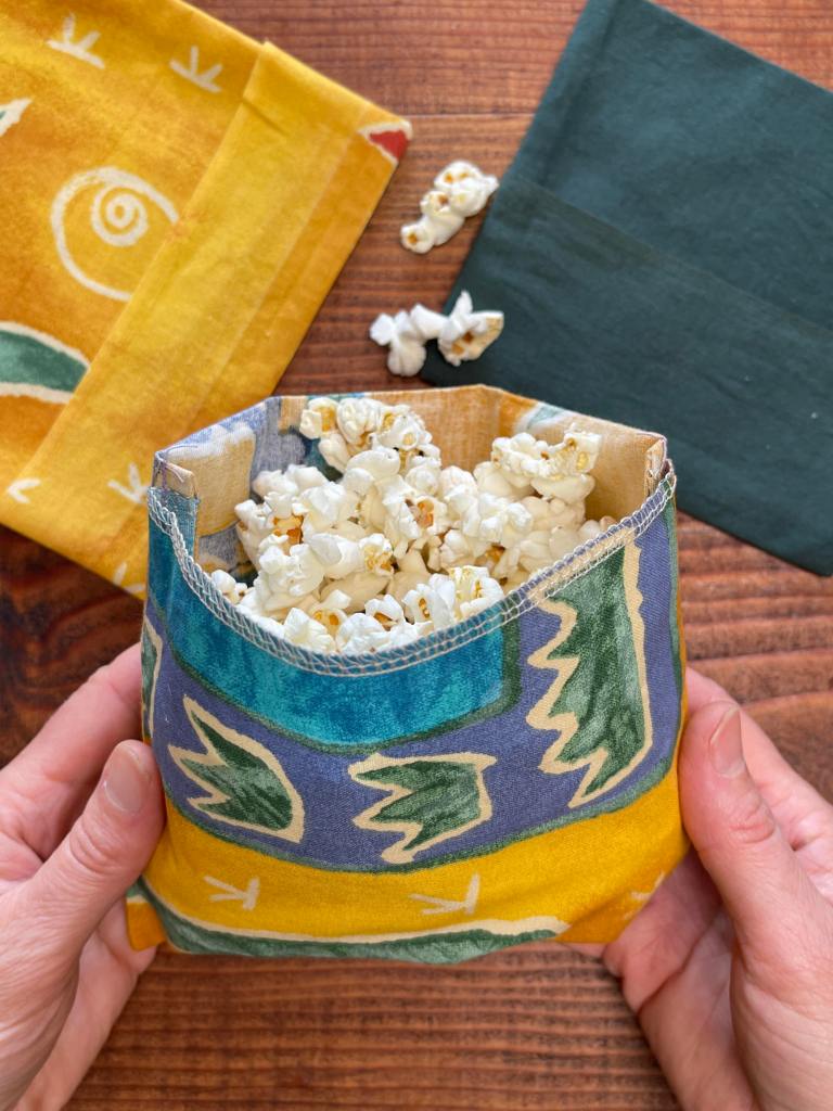 Hands hold a homemade yellow, blue and green reusable cloth snack bag filled with popcorn. Some popcorn has spilled out. An empty yellow snack bag sits to the left. On the right is an empty dark green snack bag. Everything sits on a dark wooden table.