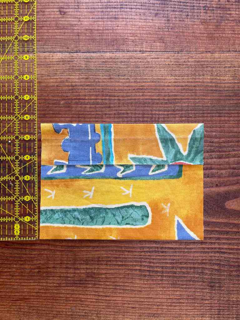 A rectangular piece of yellow, blue and green fabric is folded twice, once at the bottom and once at the top. The top overlaps. It sits on a dark wood table with a ruler next to it. The sewn snack bag will be a little over 5.5 inches high.
