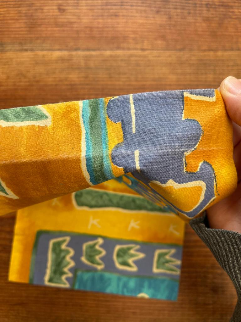 A hand holds up the selvedge edge of a piece of yellow fabric with blue and green accents