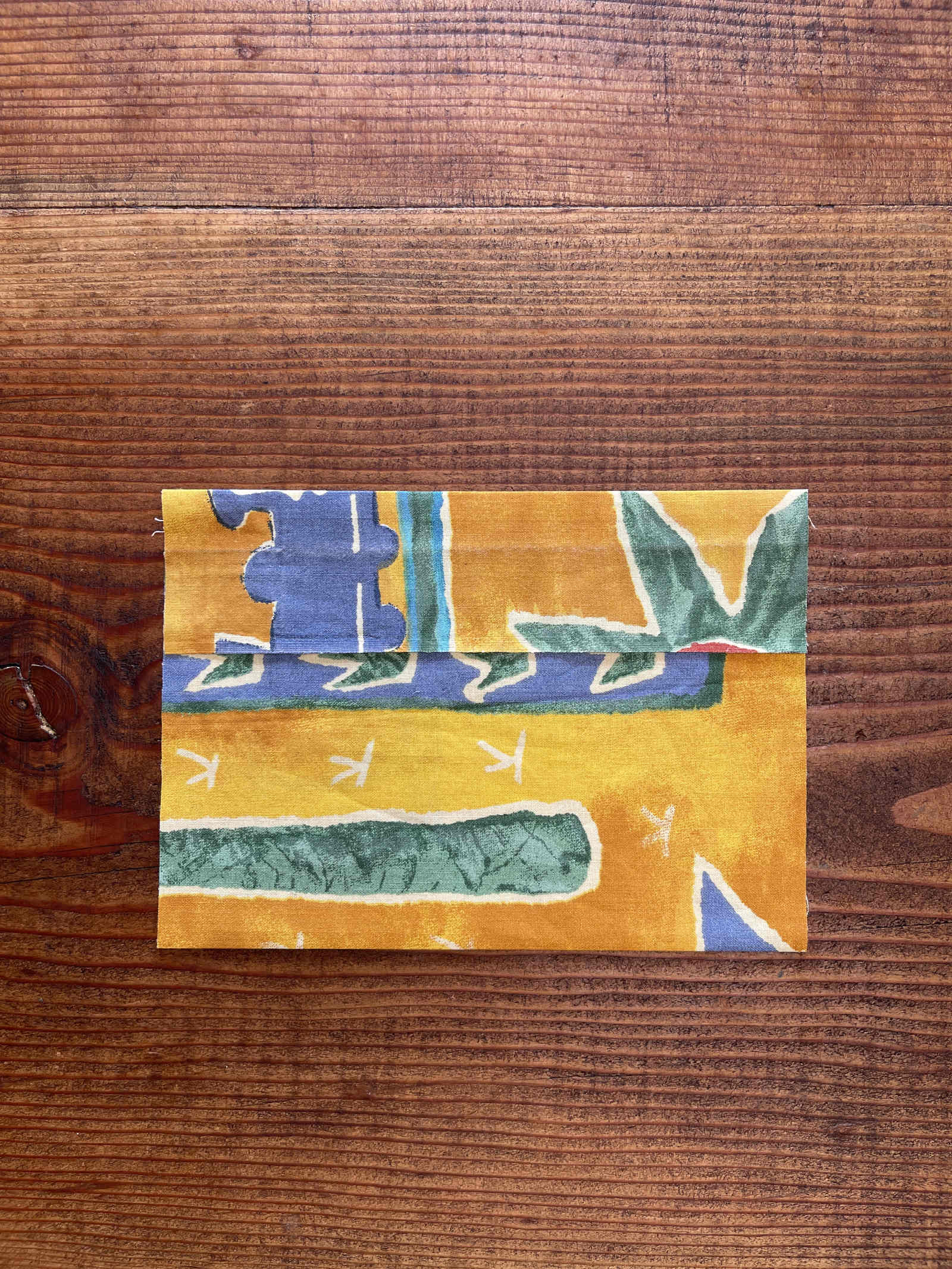A rectangular piece of yellow, blue and green fabric is folded twice, once at the bottom and once at the top. It sits on a dark wooden table.