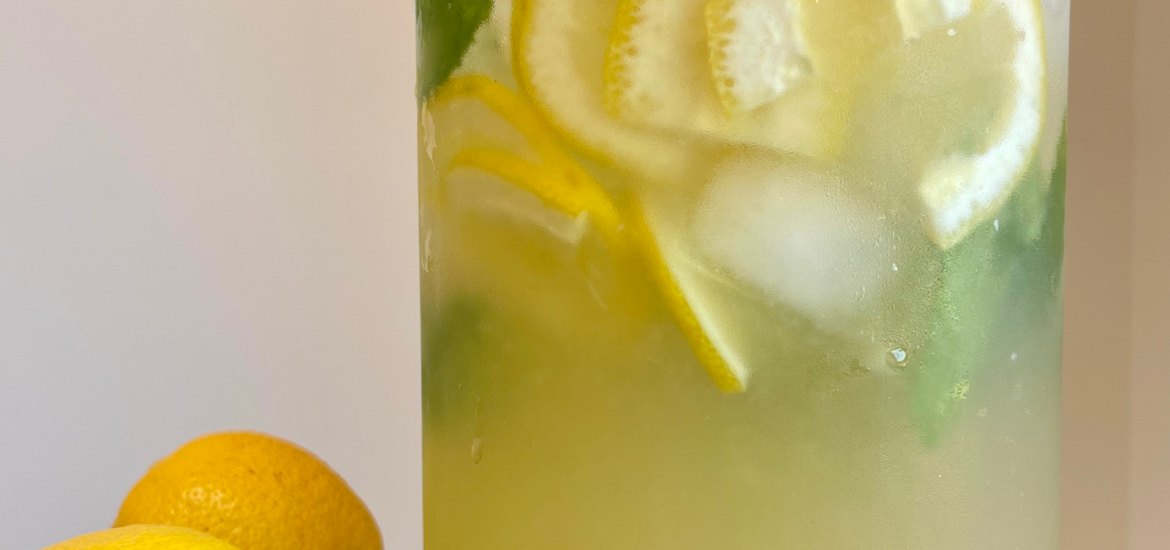 A large glass jar of lemonade with ice, lemon slices and mint. The jar sits on a wooden tabletop next to two fresh lemons..
