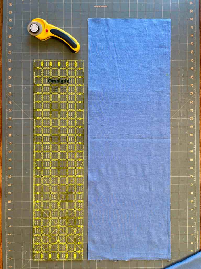 A long piece of cut blue fabric sits on a grey cutting board. Next to it is a wide rule and a rotary cutter.